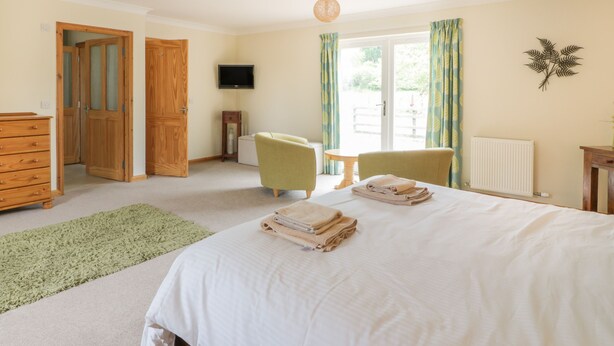A bedroom with a bed, two chairs, a dresser, a television, and a window with curtains. - Lon Cottage (Perth)