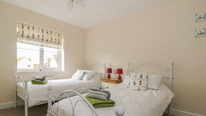 A bedroom with two single beds, each with a metal frame and white bedding. There is a window with patterned curtains, a bedside table with two red lamps, and a small shelf with decorative items.