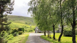 A rural pathway leading to a stone building with a blue door, surrounded by lush greenery and trees. - Stabal Cottage (Dolgellau)