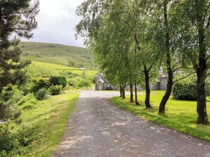 A rural pathway leading to a stone building with a blue door, surrounded by lush greenery and trees.
