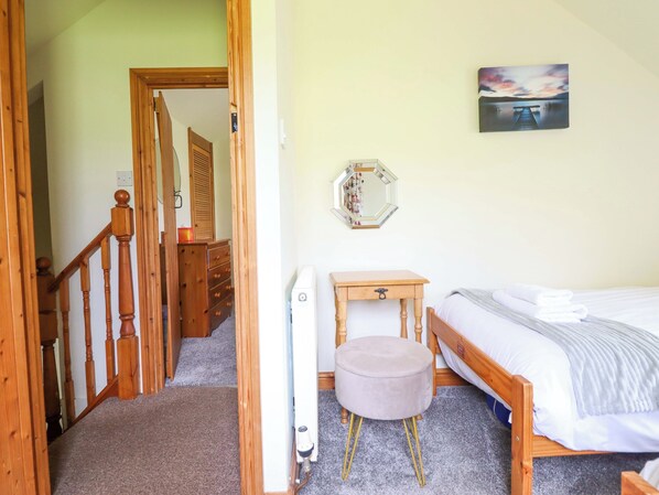 A bedroom with a bed, a wooden dresser, a round ottoman, a bedside table, a mirror, and a painting on the wall. - Stabal Cottage (Dolgellau)