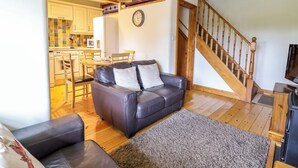 A living room with a dark leather sofa, a wooden staircase, and a kitchen in the background. - Stabal Cottage (Dolgellau)