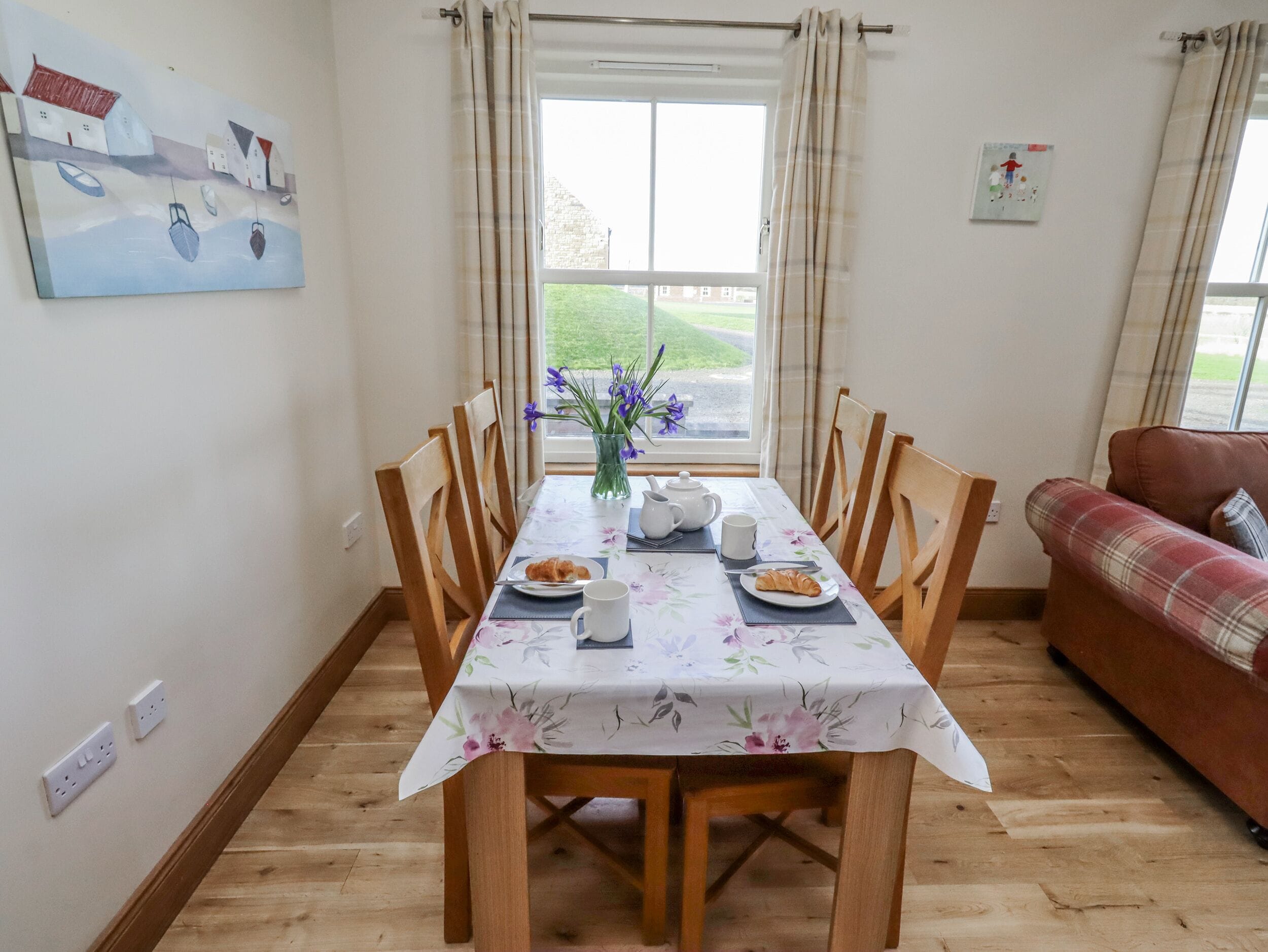 A dining area with a table set for two, a vase with flowers, and a painting on the wall.