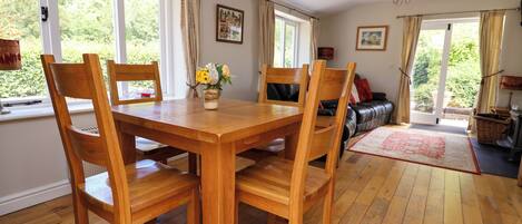A dining area with a wooden table and chairs, a vase with flowers, and a view of greenery through the windows.
