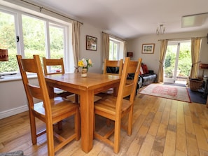 A dining area with a wooden table and chairs, a vase with flowers, and a view of greenery through the windows.