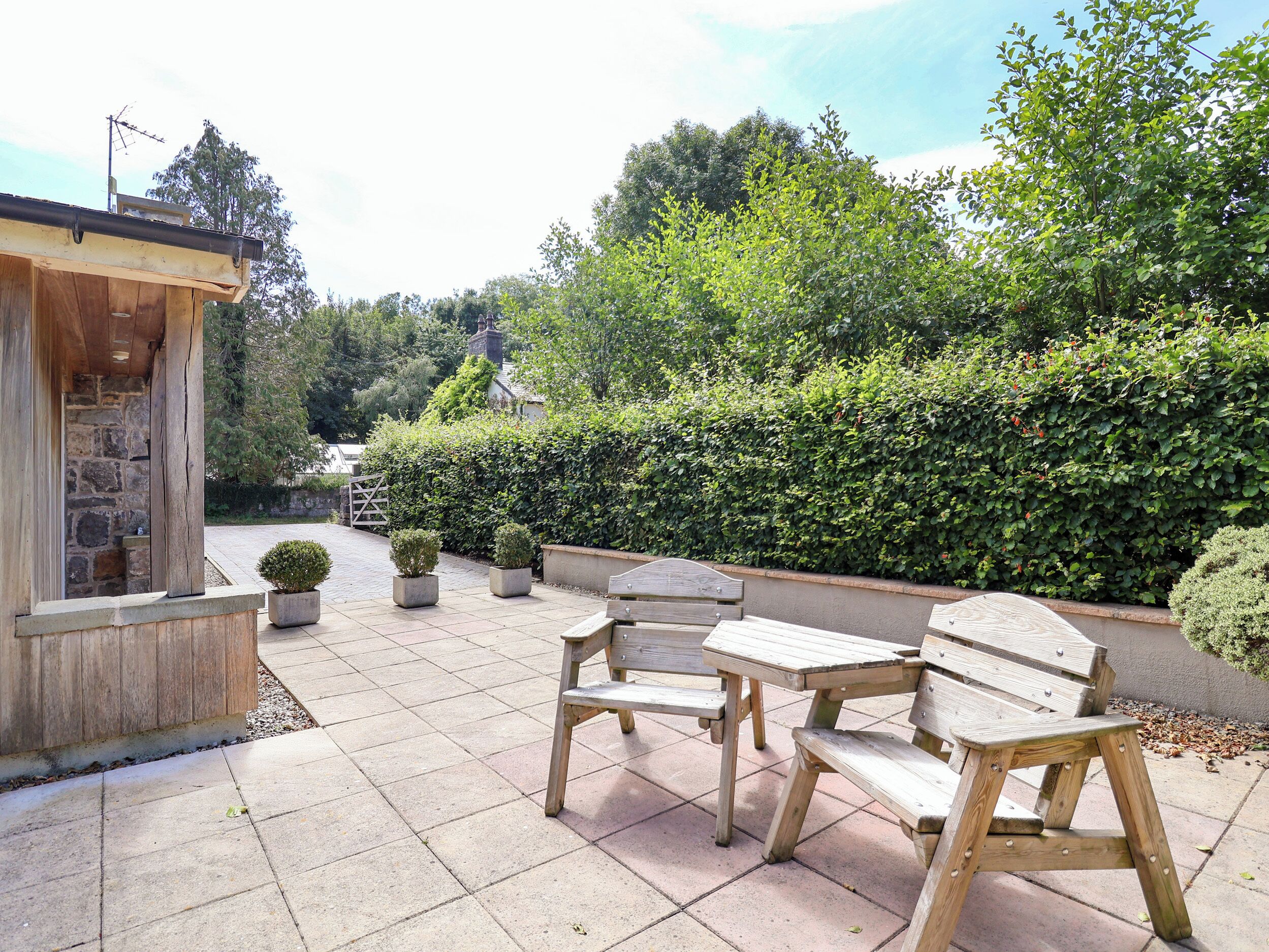 An outdoor patio with a wooden table and chairs, surrounded by potted plants and a stone wall.