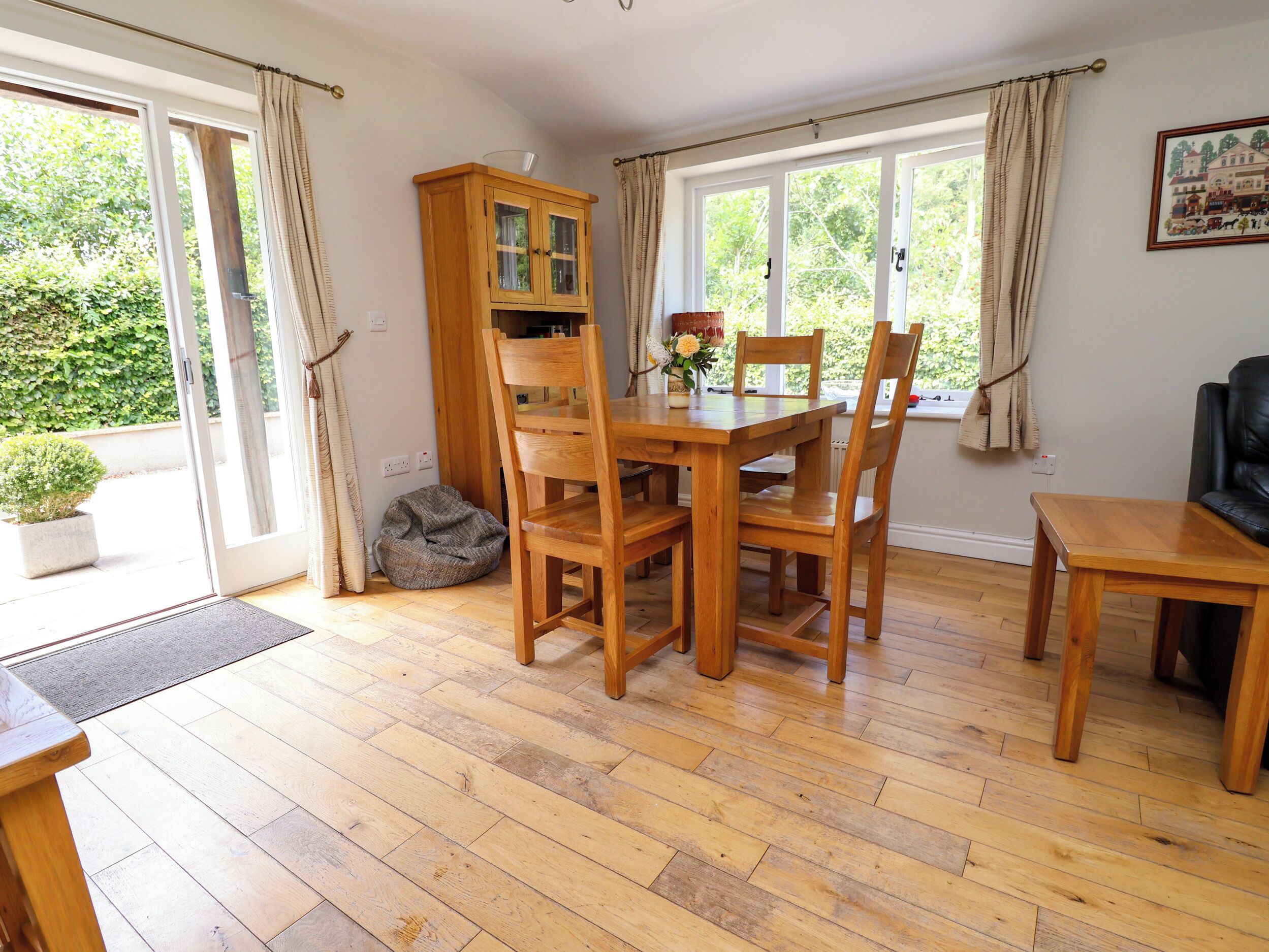 A dining area with a wooden table and chairs, a cabinet with glass doors, and a view of greenery outside.
