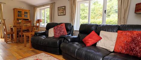 A living room with a leather sofa, a wooden dining table, and chairs. There are large windows with curtains, a chandelier, and a framed picture on the wall.