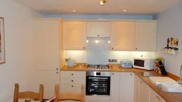 A compact kitchen with white cabinets, a gas stove, a microwave, and a sink. There is a wooden chair and a table with a bottle and a box on it.