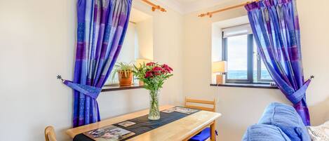 A cozy room with a wooden dining table, blue chairs, and a blue sofa. A vase with flowers is on the table, and curtains are drawn on the windows.