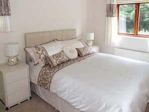 A neatly made bed with a white duvet and patterned pillows, a bedside table with a lamp, and a window with curtains.