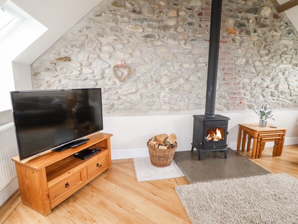 A cozy living room with a stone wall, a fireplace, a TV on a wooden stand, and a small table with a vase. - Yr Hen Festri (Caernarfon)