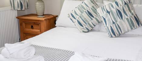 A neatly made bed with white linens and striped pillows, a wooden nightstand with a lamp, and a radiator in the background.