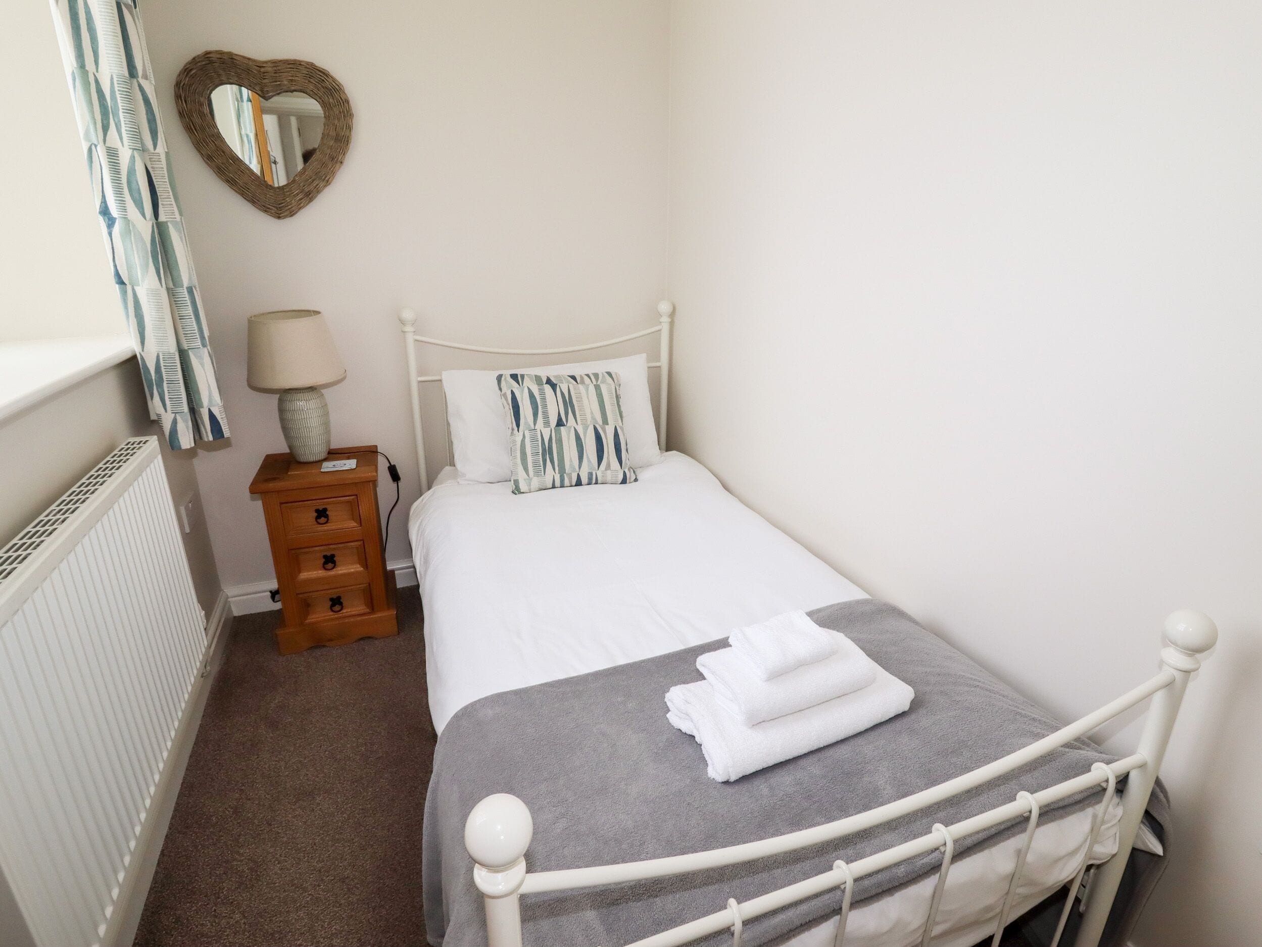 A neatly arranged bedroom with a single bed, a bedside table with a lamp, a dresser, a radiator, and a heart-shaped mirror on the wall.