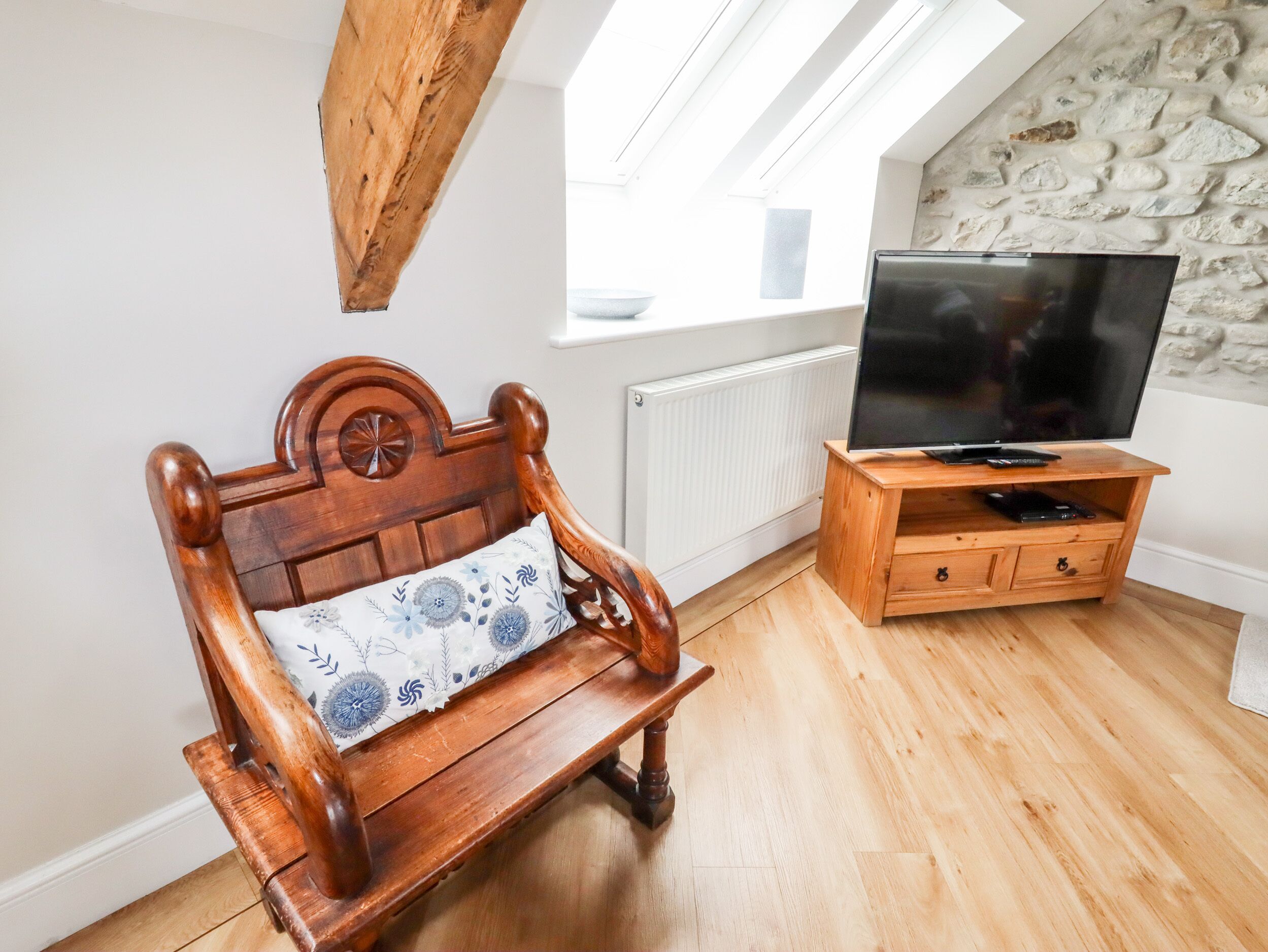 A wooden bench with a floral cushion, a television on a wooden stand, and a stone wall in a room with wooden flooring.
