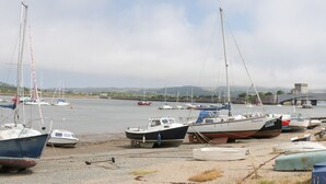 A marina with several boats docked on the shore, a bridge in the background, and a castle-like structure across the water. - Bryn Derw Holiday Cottage (Colwyn Bay)