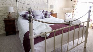 Una cama con dosel de metal blanco con una manta roja, una mesita de noche de madera con una lámpara y una cortina estampada con flores.