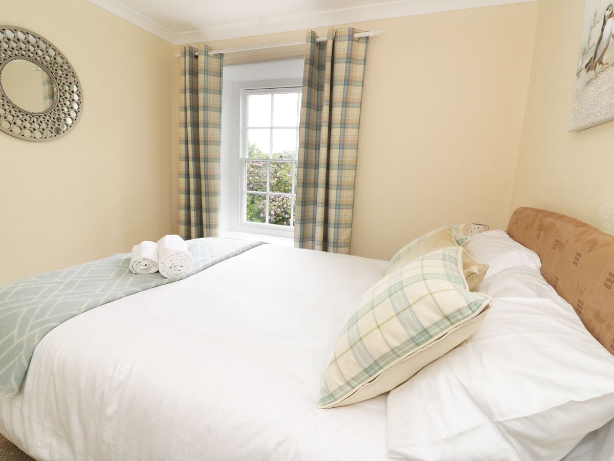 A neatly made bed with a plaid pillow and a rolled towel, a window with curtains, and a wall-mounted mirror.