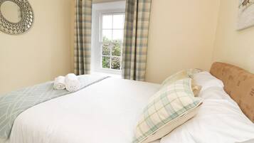 A neatly made bed with a plaid pillow and a rolled towel, a window with curtains, and a wall-mounted mirror.