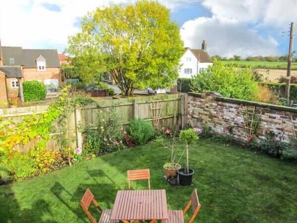A backyard with a wooden table and chairs, a brick wall, and a variety of plants and flowers. - Old Church Coach House (Stoke-on-Trent)