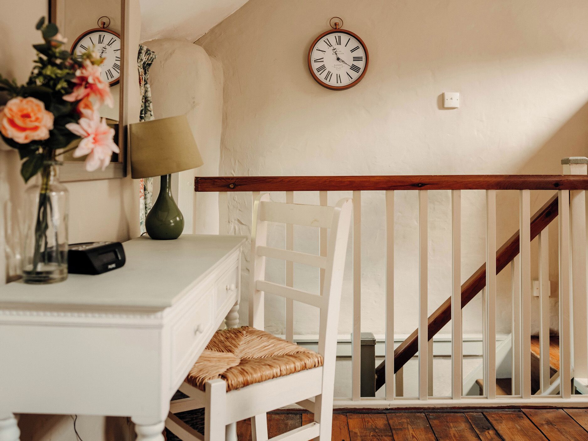 A white chair with a woven seat, a white dresser with a lamp, and two wall clocks.