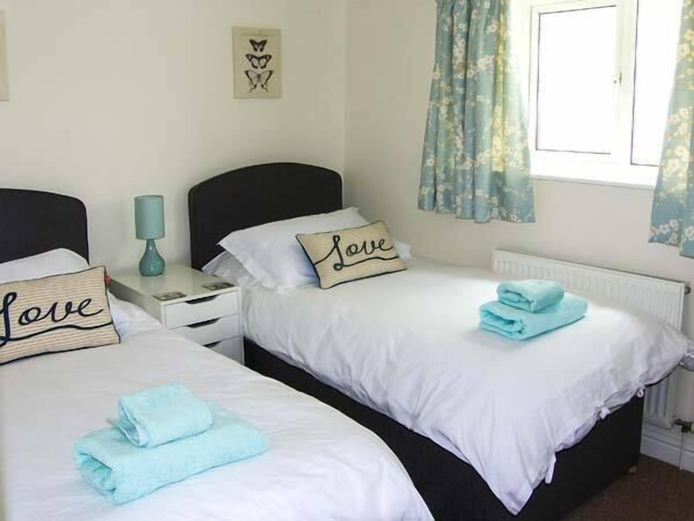 Une chambre avec deux lits, chacun orné d’un coussin sur lequel est brodé le mot « Amour ». Il y a une fenêtre avec des rideaux, une table de chevet avec une lampe et un radiateur.