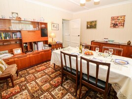 A dining area with a table set for four, surrounded by wooden chairs. A bookshelf filled with books and decorative items is to the left. The room has patterned carpet and a door leading to another area.