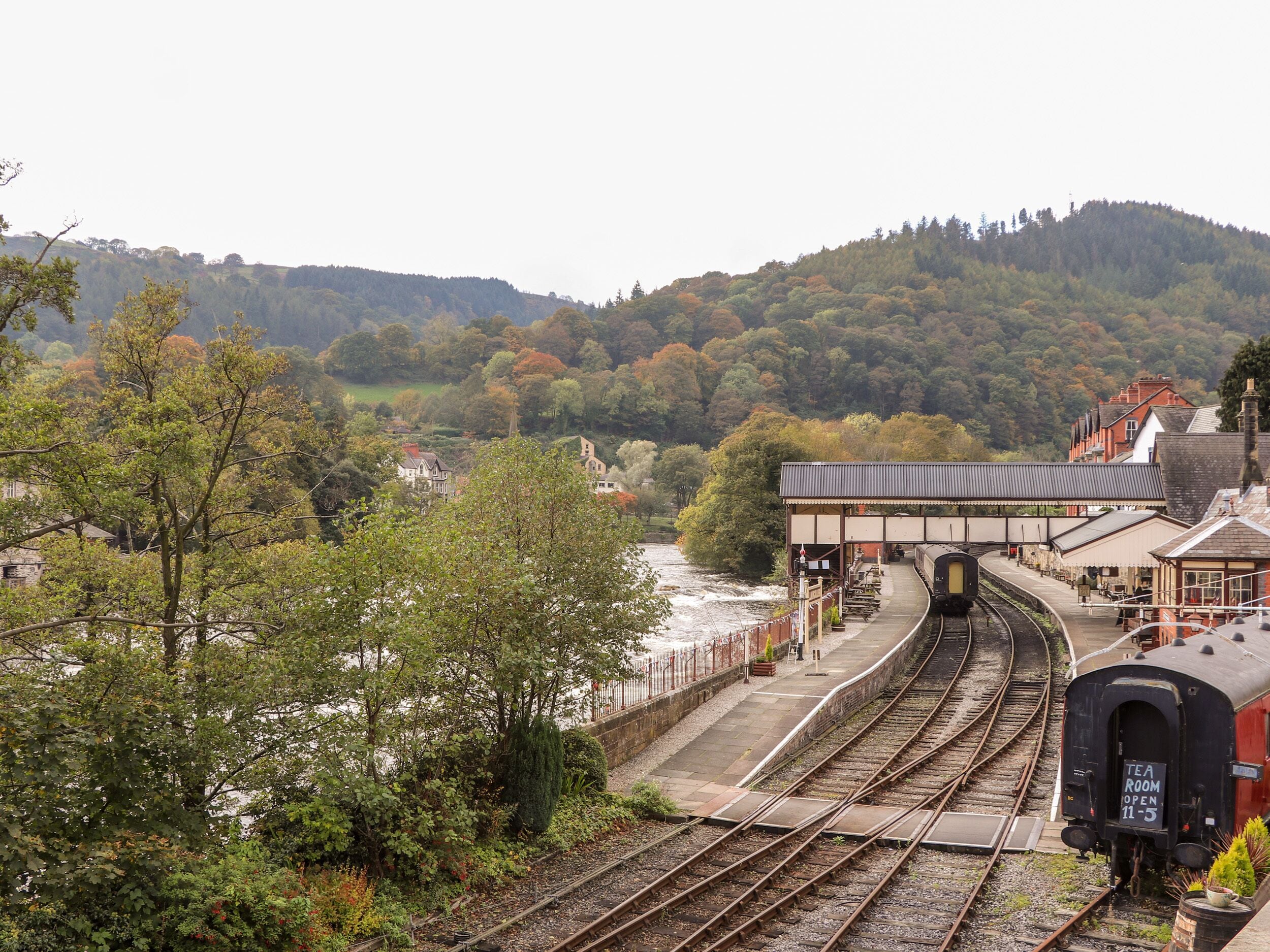 A train station with multiple tracks, a bridge over a river, and a backdrop of hills.