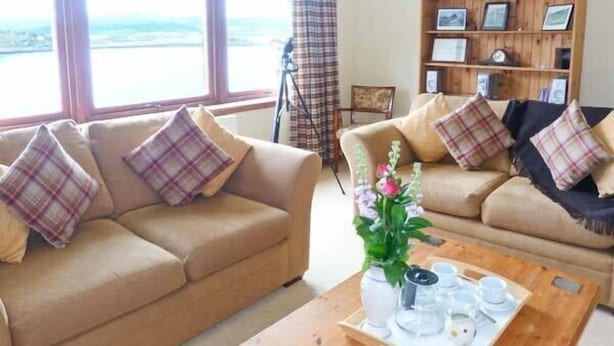 A living room with two beige sofas, each with plaid cushions, a wooden coffee table with a tray of white cups and saucers, and a bookshelf with framed pictures. - Mackenzies Peak (Sconser)