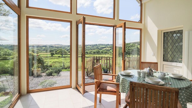 A dining area with a table set for four, large windows offering a view of a green landscape, and a wooden chair in the foreground. - Llety'r Wennol (Llanwrda)