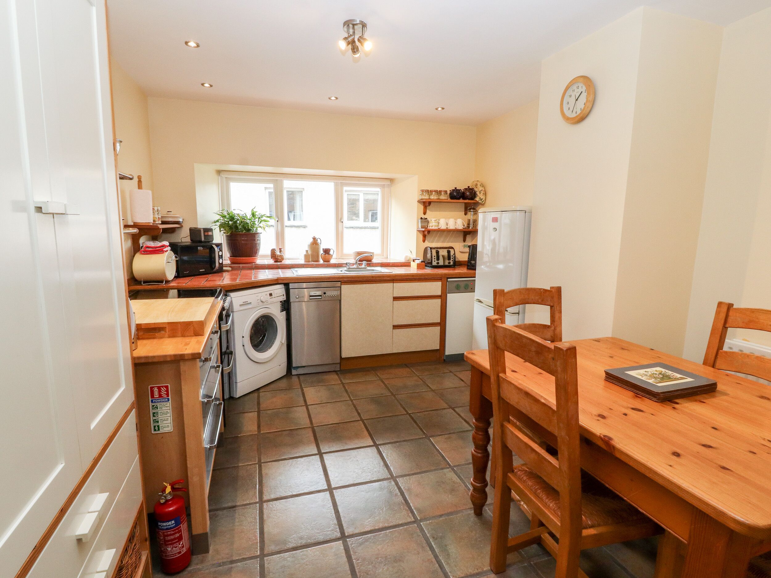 A compact kitchen with a washing machine, a dishwasher, and a refrigerator. There's a dining table with chairs and a fire extinguisher on the floor.