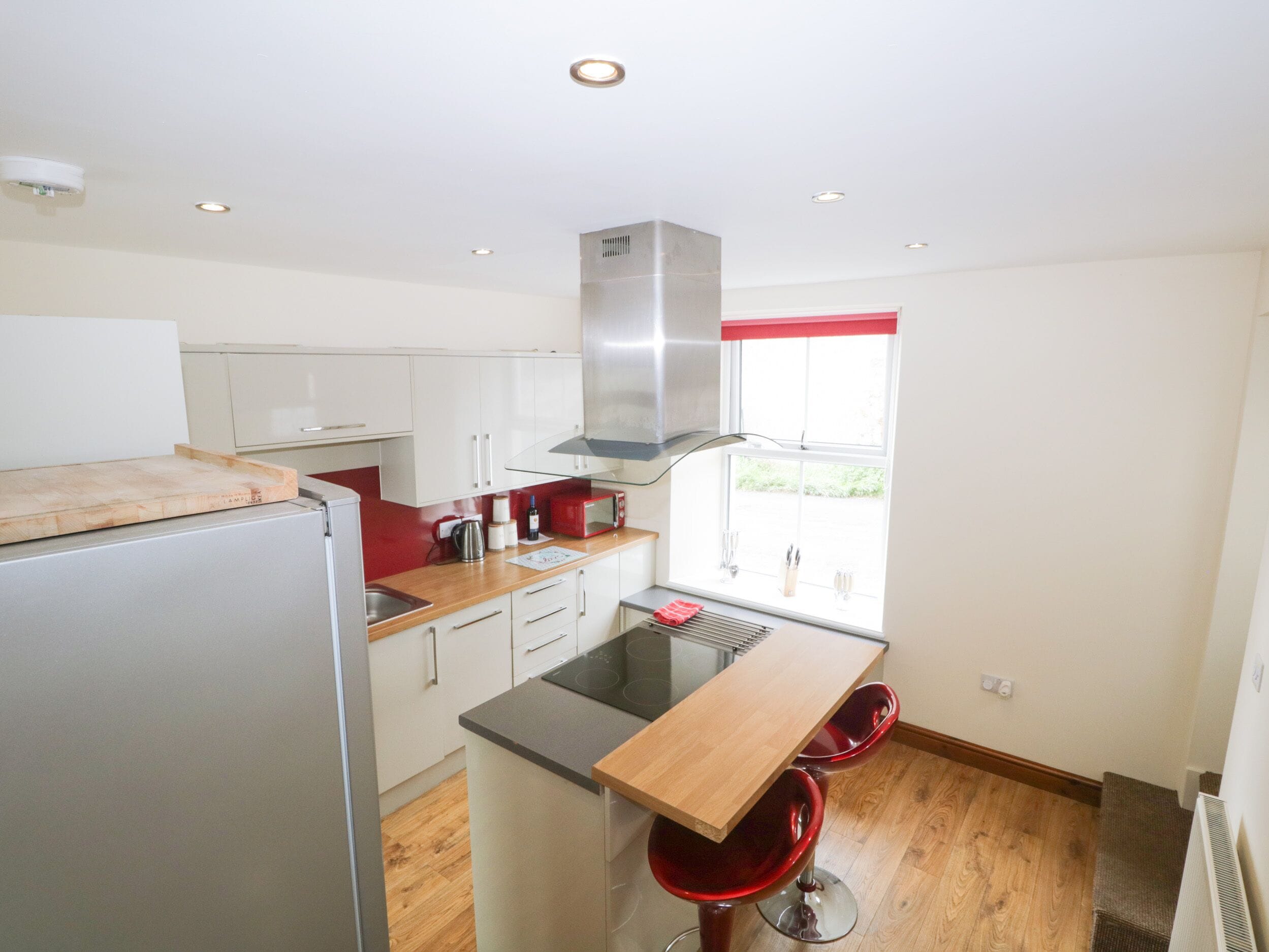 Una cocina compacta con armarios blancos, una campana extractora de acero inoxidable, una encimera de madera y una ventana con cortina roja.