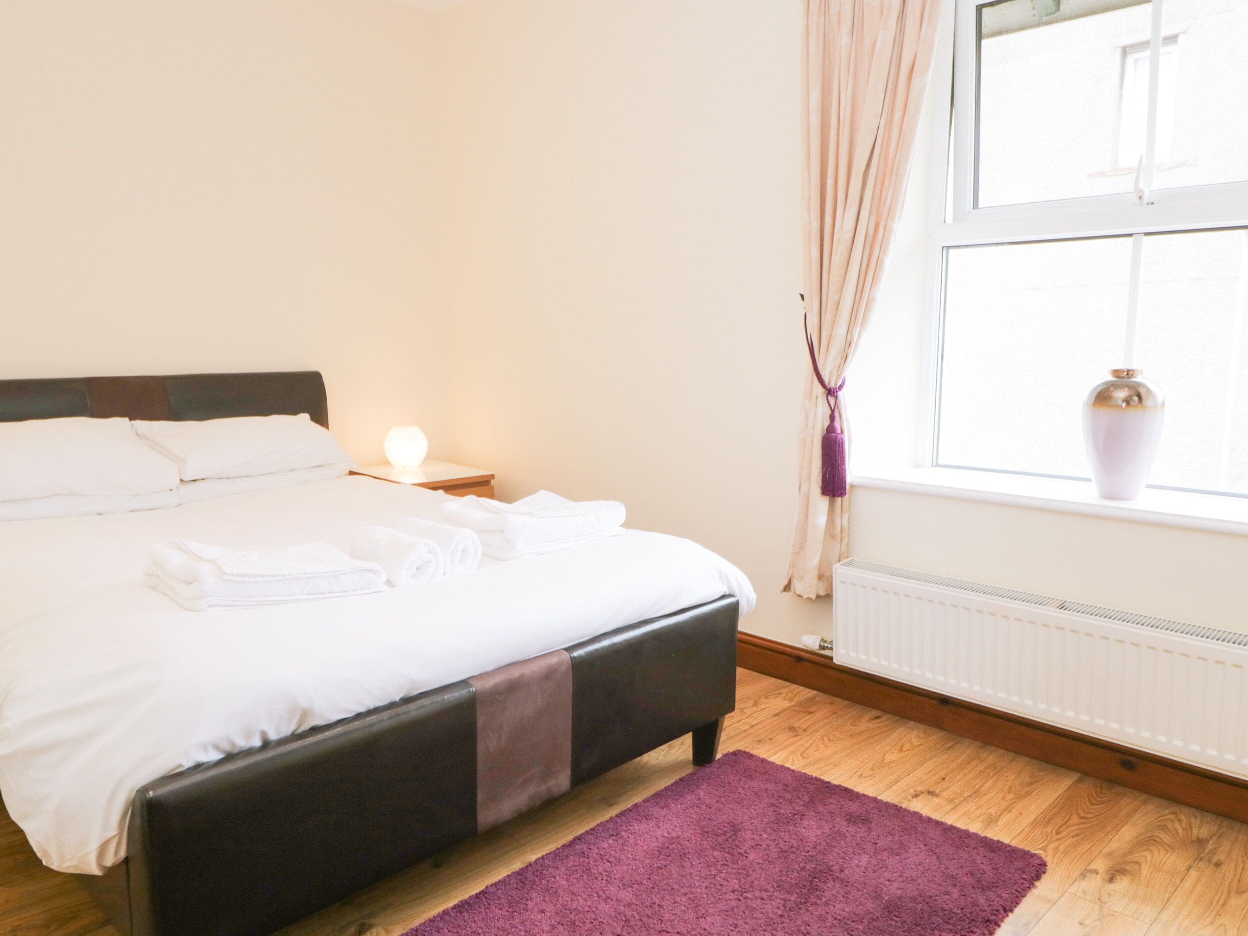 A neatly made bed with white linens and a purple throw blanket, a bedside lamp, a window with curtains, and a radiator.