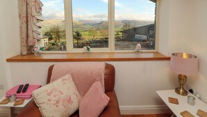 A room with a large window offering a view of a rural landscape, a wooden bench with cushions, a small table with a lamp, and a pink remote control on a side table. - Hawkrigg Cottage (Carnforth)