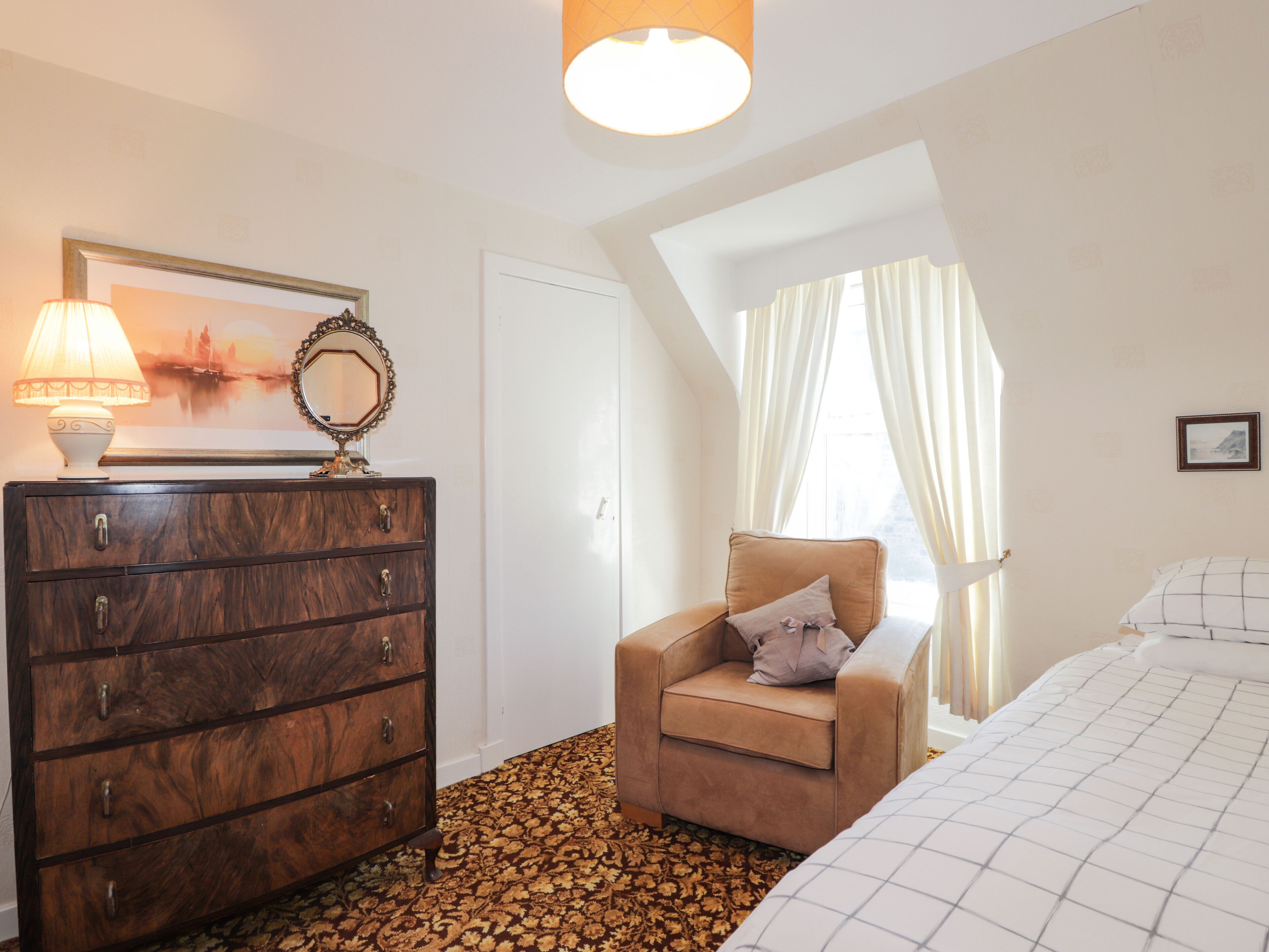 A bedroom with a wooden dresser, a chair, a bed, a window with curtains, and a lamp.