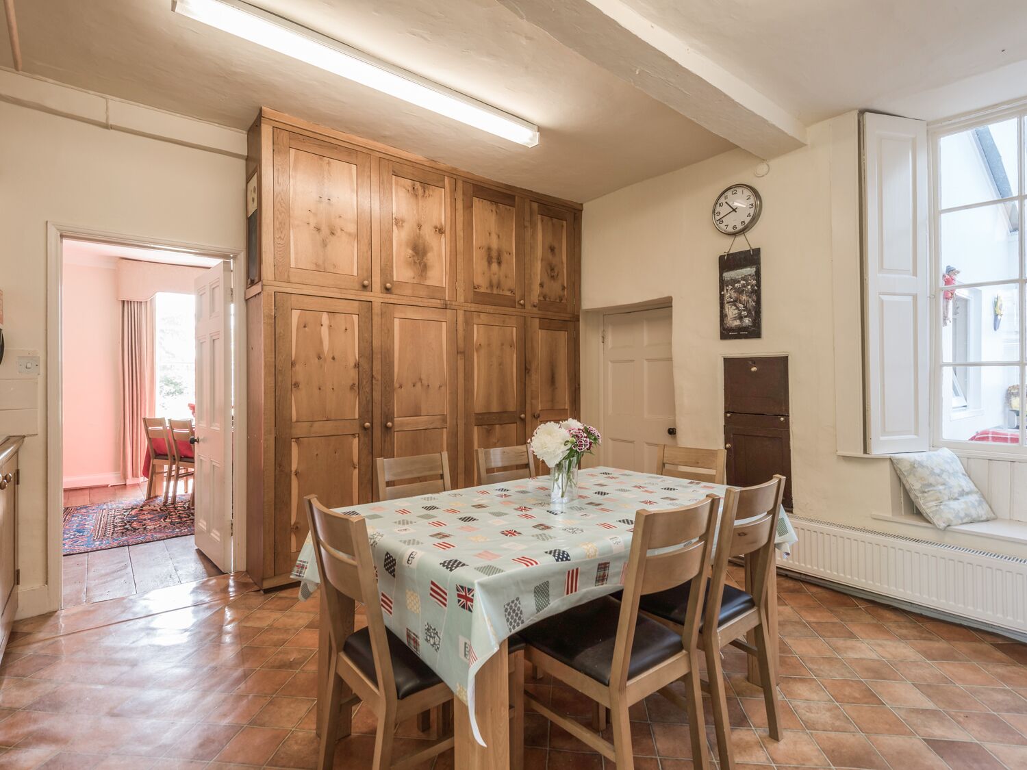 Uma sala de jantar com uma mesa posta para quatro pessoas, armários de madeira, um relógio na parede e uma janela com vista.