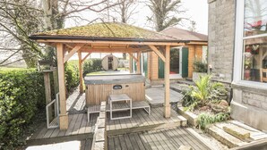 A wooden deck with a hot tub and seating area, surrounded by greenery and a stone building. - Eden Lodge (Ulverston)