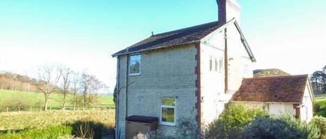 A single-story stone house with a chimney, surrounded by a rural landscape.