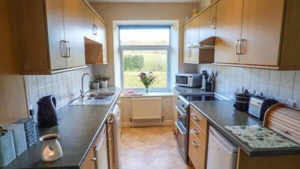 A compact kitchen with wooden cabinets, a built-in microwave, and a window providing a view of the outdoors. - Pensychnant Cottage (Conwy)