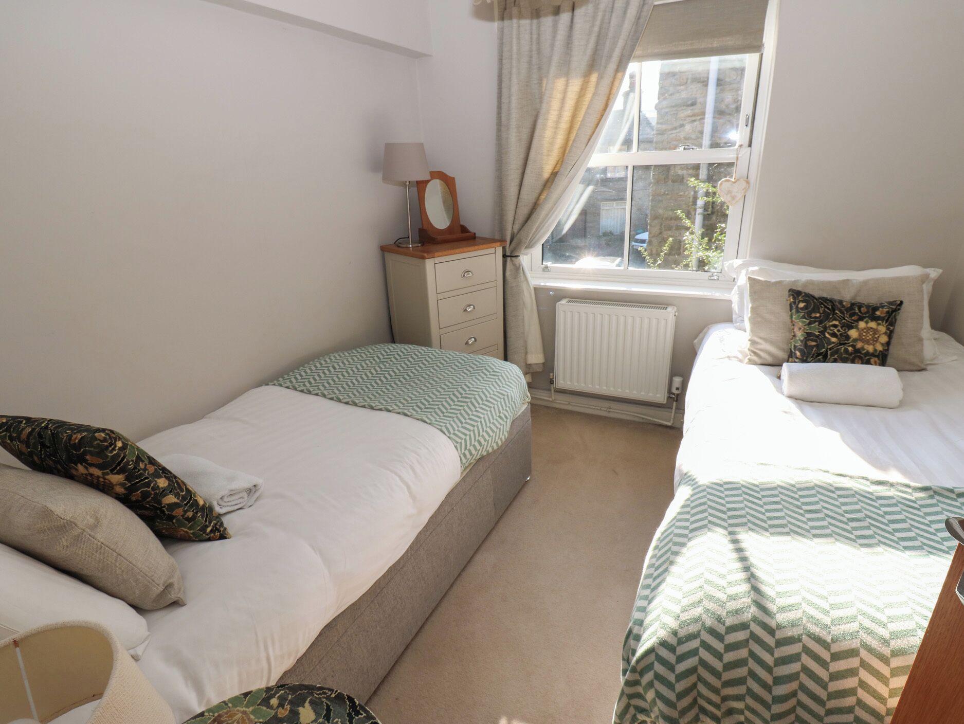 A bedroom with two beds, a bedside table with a lamp, a radiator, and a window with curtains.