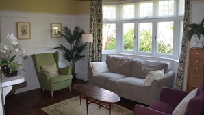 Un salon avec un fauteuil vert, un canapé gris, une table basse en bois et une grande fenêtre avec des rideaux.