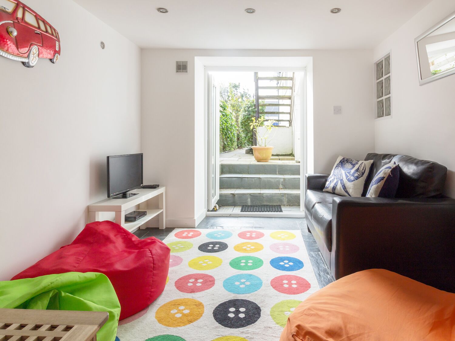 Een woonkamer met een televisie, een bank, een kleurrijk knopenkleed en een rode puf.