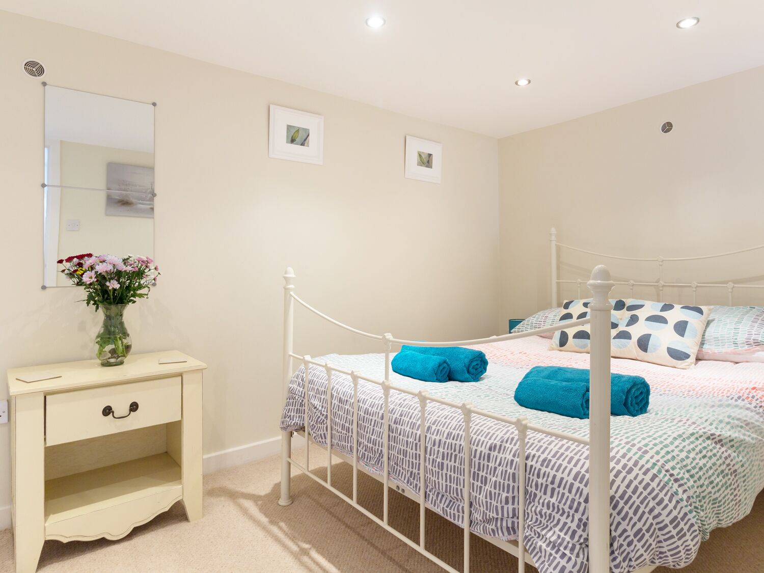 A bedroom with a white metal bed frame, a bedside table with a vase of flowers, and two framed pictures on the wall.
