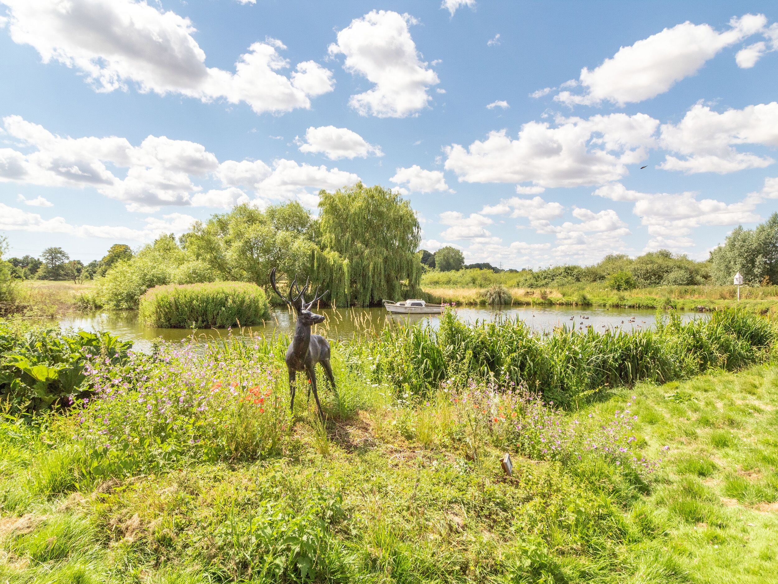 A serene riverside setting with a deer statue, lush greenery, and a weeping willow tree.