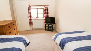A bedroom with two beds, a wooden dresser, a television on a stand, and a window with curtains. - Sunnymead Cottage (Beaworthy)