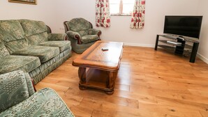 A living room with a sofa set, a wooden coffee table, a television on a stand, and a window with floral curtains. - Sunnymead Cottage (Beaworthy)