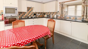 A compact kitchen with white cabinets, a microwave, a toaster, and a coffee maker. There is a dining table with a red polka dot tablecloth and two wooden chairs. - Sunnymead Cottage (Beaworthy)