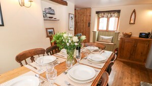 A dining area with a wooden table set for four, surrounded by chairs. There is a vase with flowers on the table, and a wooden cabinet with a radio on top.