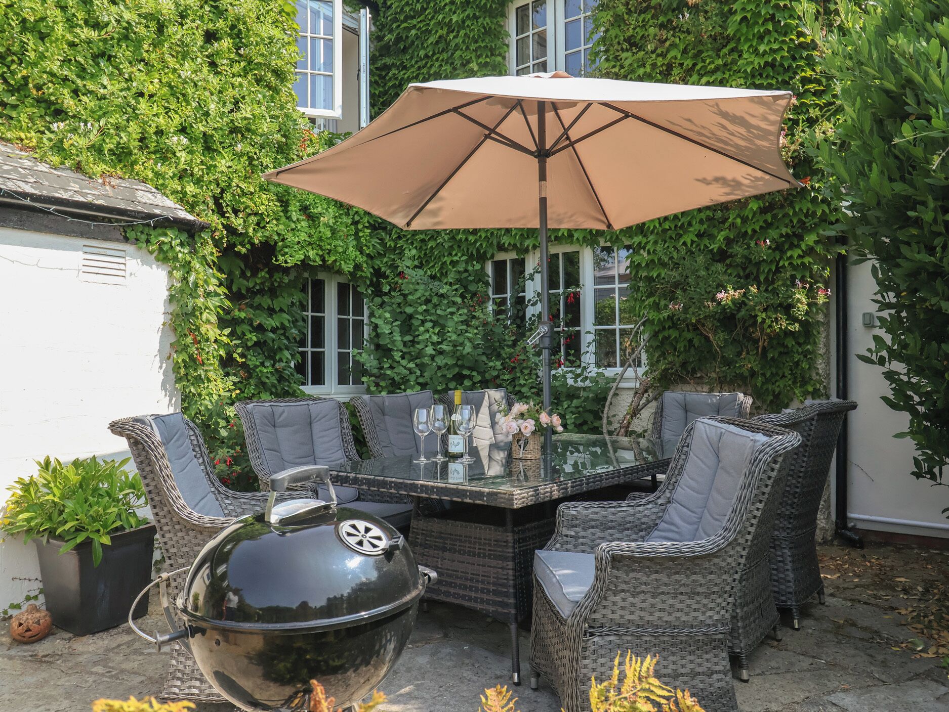 An outdoor patio with a glass-top table, wicker chairs, a barbecue grill, and a large umbrella.