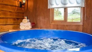 A wooden cabin interior with a hot tub, a window with a white blind, and a wall-mounted lamp.